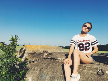 Portrait of young woman wearing sunglasses while sitting on footpath against clear blue sky during sunny day