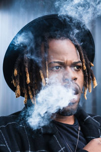 Side view of ethnic back guy with dreadlocks and hat in dense cloud of fume smoking on street in city