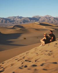 Couple sitting on sand dune in desert against sky