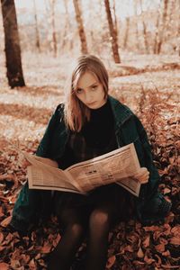 Portrait of young woman holding newspaper while sitting on field during autumn