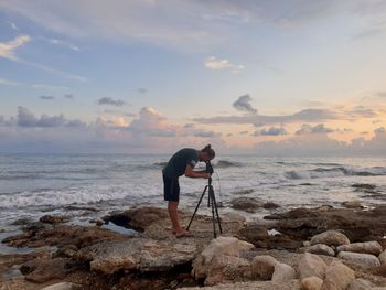 Man photographing on rock at beach against sky during sunset