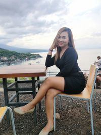 Young woman sitting on bench against sky