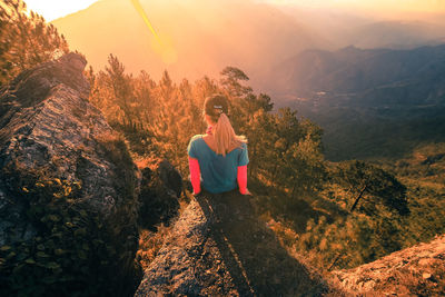 Rear view of teenager sitting on cliff against mountains