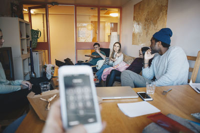 Cropped hand woman holding mobile phone against friends in college dorm room