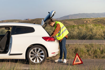 Young man on a yellow vest trying to find something to fix the broken car on the side of the road 