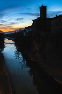 Silhouette buildings by river against sky at sunset