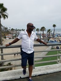 Mature man wearing sunglasses standing by railing against beach