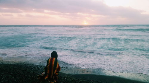 Person covered blanket sitting at sea shore against sky during sunset