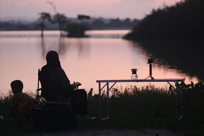 Rear view of silhouette men sitting by lake against sky during sunset