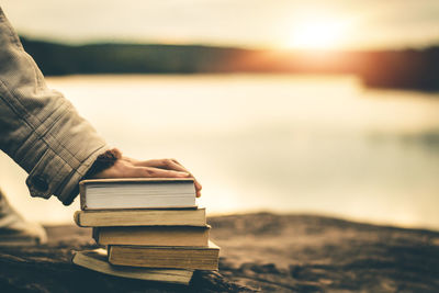 Person holding book at beach against sky during sunset