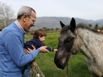 Side view of happy grandfather assisting grandson in touching horse at farm