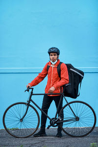 Portrait of delivery boy standing by bicycle against wall