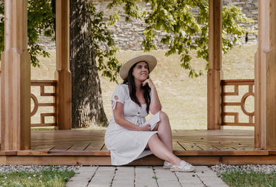 Portrait of beautiful young woman wearing white dress sitting under wooden pavilion in park.