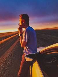 Side view of man smoking cigarette while standing by car on road against sky during sunset