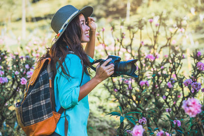 Side view of smiling young woman holding camera while standing amidst flowers on land