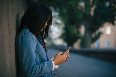 Side view of teenage girl using smart phone while standing against wall