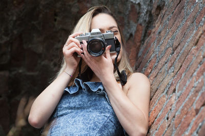 Low angle view of young woman photographing while standing by brick wall