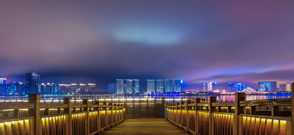 Illuminated bridge by buildings against sky at night