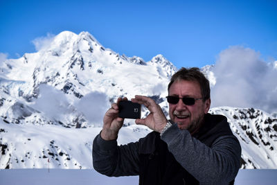Smiling mature man photographing with mobile phone while standing against snow covered mountains