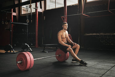 Full length of muscular man sitting on weights in gym