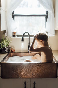 Shirtless boy taking bath while sitting in kitchen sink at home