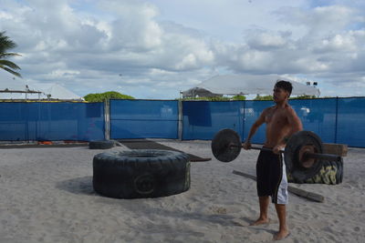 Full length of man lifting barbell while standing on sand against cloudy sky