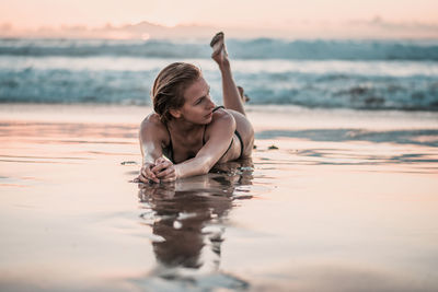 Full length of seductive woman lying on shore at beach against sky during sunset