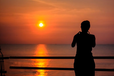 Rear view of silhouette woman standing by railing against sea and orange sky