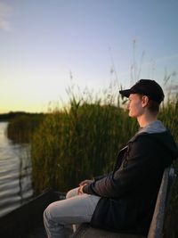 Young man looking away while sitting on lake against sky