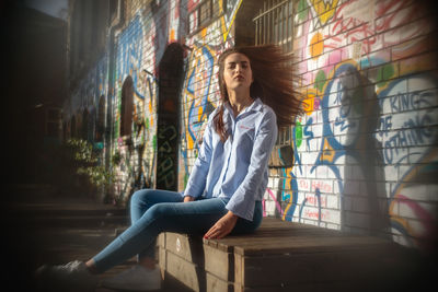 Portrait of confident young woman sitting on wood against graffiti wall in city