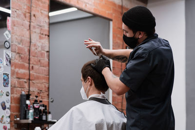 Barber in protective mask and latex glove styling adult man in a barbershop salon. new normal