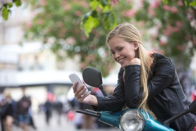 Smiling woman using mobile phone while leaning on motor scooter