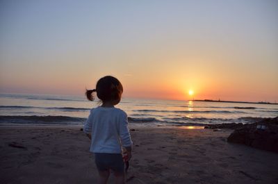 Rear view of girl standing at beach against sky during sunset