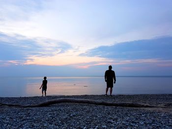 Rear vie of father with son standing on shore at beach during sunset