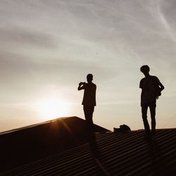 Silhouette friends on house roof against sky during sunset