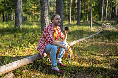 Man camper hiker sitting on log in forest enjoying drinking tea from thermos having break.