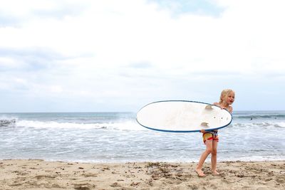 Full length of girls holding surfboard while standing on shore at beach against sky