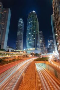 Light trails on city street by buildings against sky at night