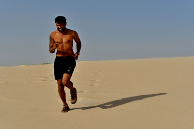 Full length of shirtless young man running on sand at desert during sunny day