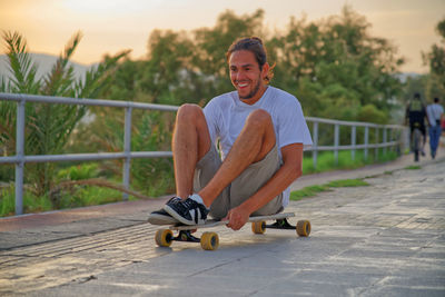 Full length of smiling young man sitting on skateboard during sunset