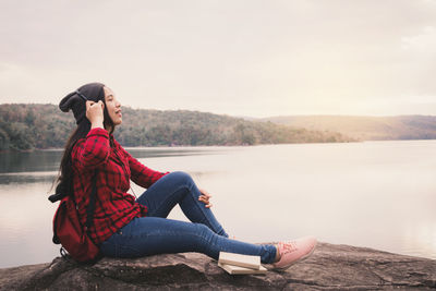 Young woman sitting on rock by lake against clear sky