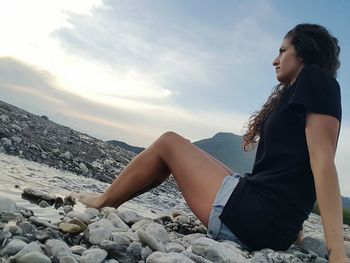 Side view of woman relaxing at beach against sky during sunset