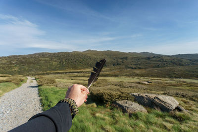 Cropped hand of man holding feather over landscape against sky