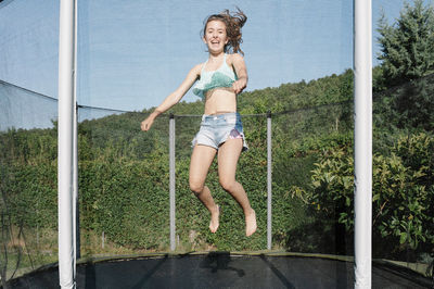 Horizontal photo of a smiling young brunette teenager jumping on a trampoline with net around on the green yard outdoors. the girl wears short jeans and a top and looks fun, active