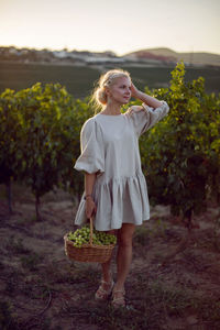 Woman with a wicker basket of green grapes stands in her vineyard at sunset