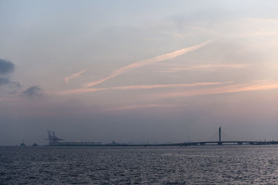 Distant view of silhouette bridge over sea against sky during sunset