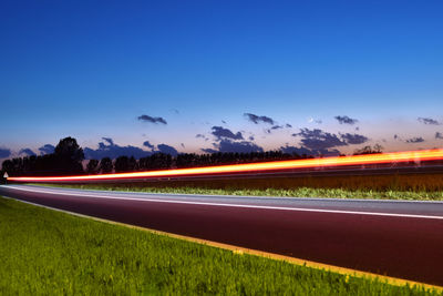 Light trails on road amidst field against blue sky