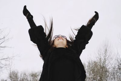 Smiling young woman standing with arms raised against clear sky