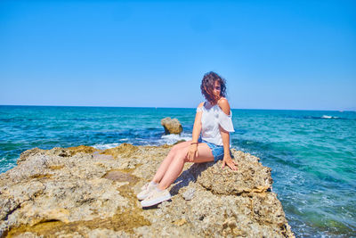 Young woman sitting on rock at beach against clear blue sky