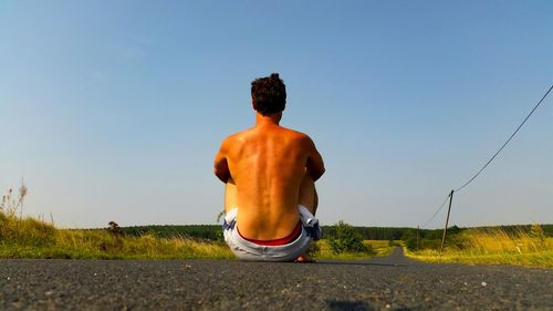 Rear view of shirtless man sitting on road against sky